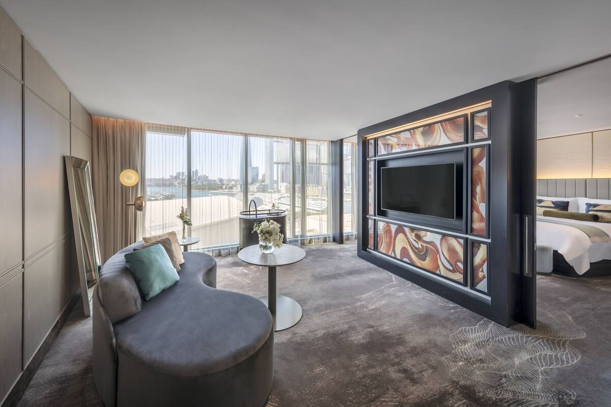 Una habitación de hotel moderna con un televisor grande, un sofá, una mesita redonda con un jarrón de flores y vistas a la ciudad.
