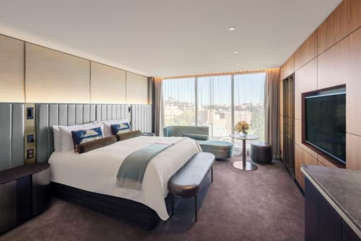 A modern hotel room with a large bed, a TV, a small round table, and a bench.