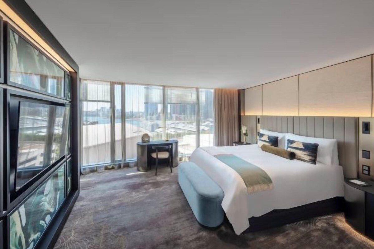 A modern hotel room with a large bed, a small desk, and a view of the city through large windows.