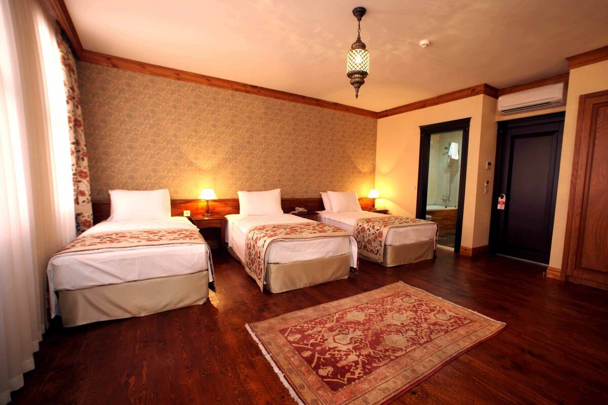 A hotel room with three single beds, a wooden floor, a patterned rug, and a hanging light fixture.