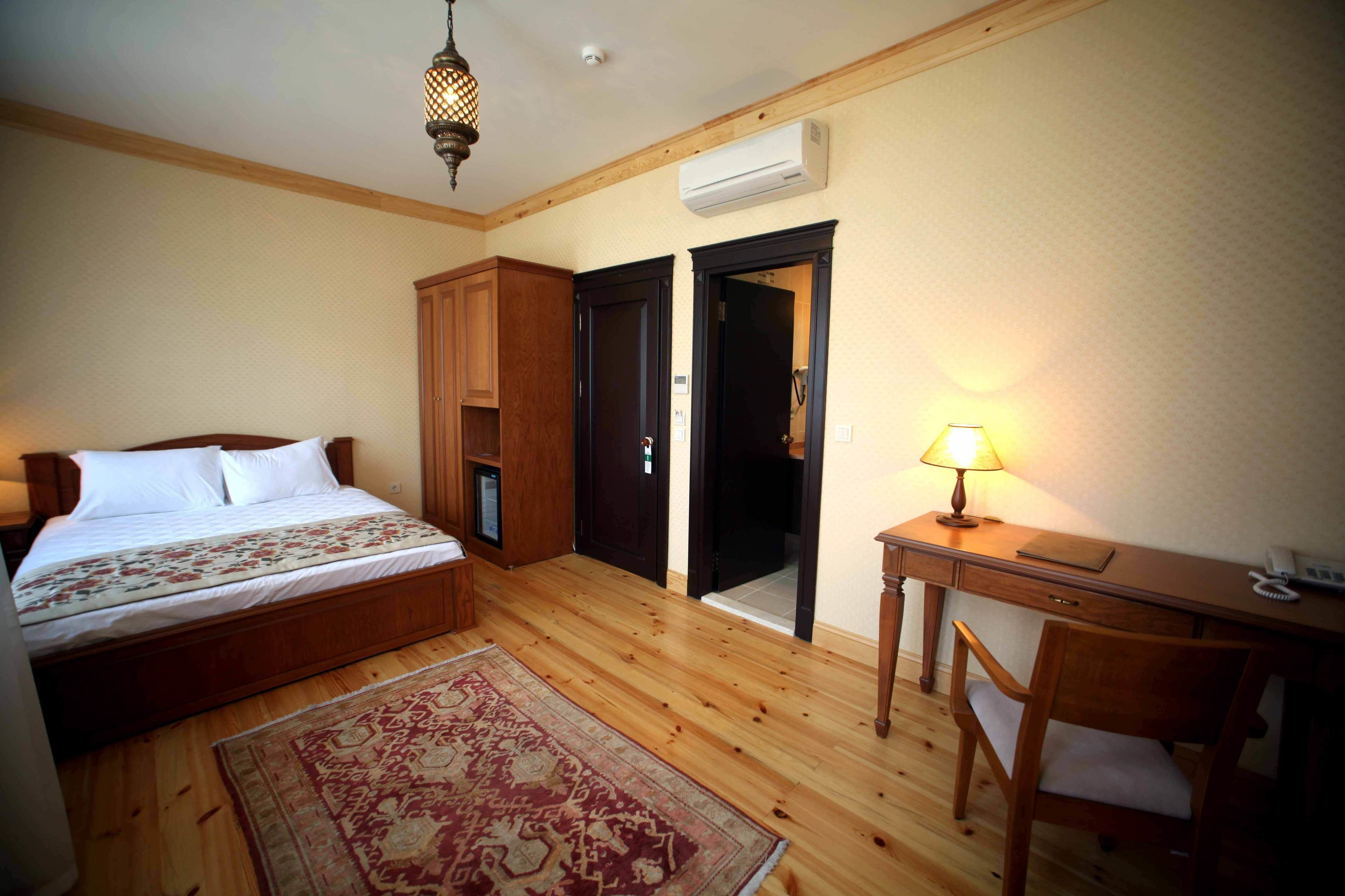 A bedroom with a wooden bed, a desk, and a chair. There is a door leading to another room and an air conditioning unit on the wall.