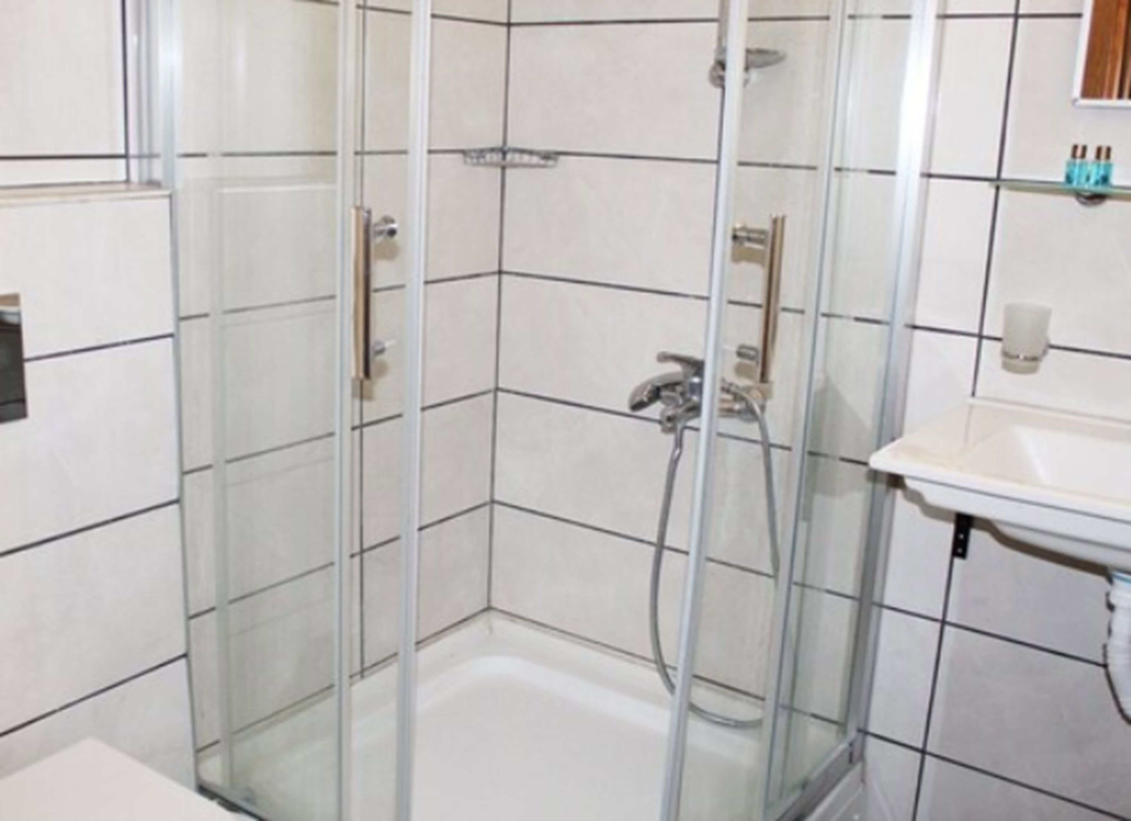 Una ducha en esquina con puertas de vidrio, pared de azulejos blancos y lavamanos con espejo arriba.