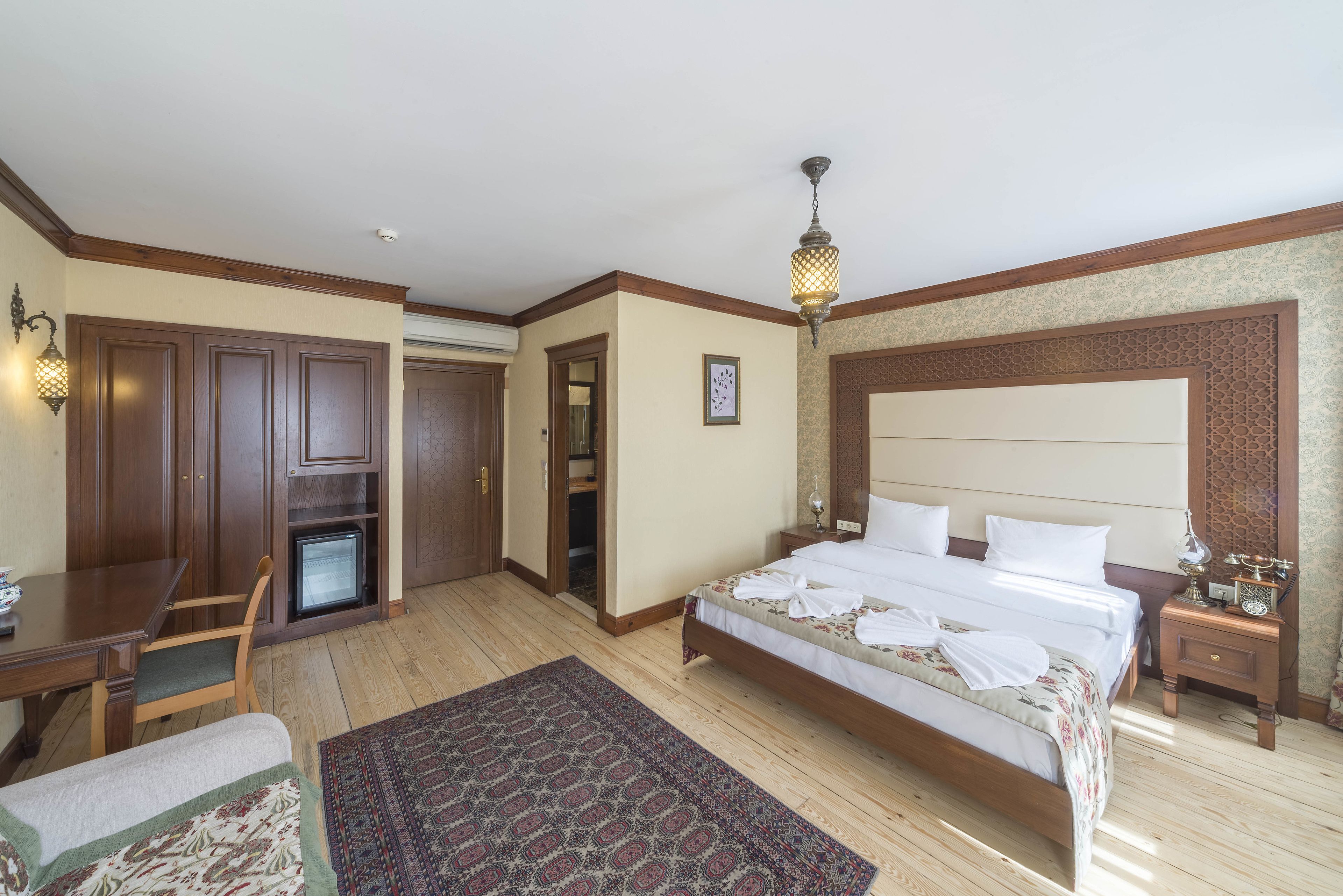 A hotel room with a large bed, a desk, a chair, a small refrigerator, and a decorative rug.