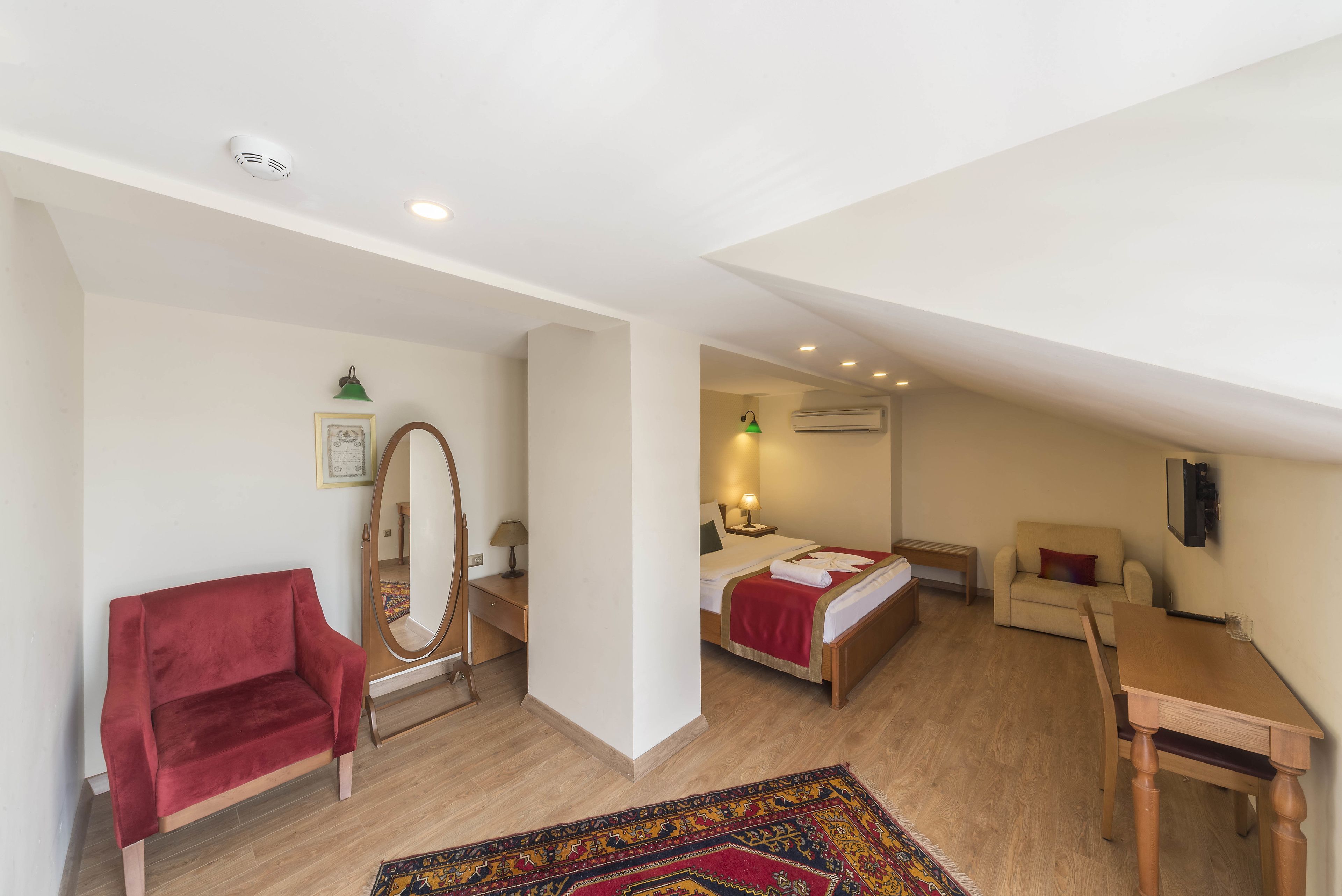 A bedroom with a bed, a red chair, a wooden table, a mirror, and a small desk.