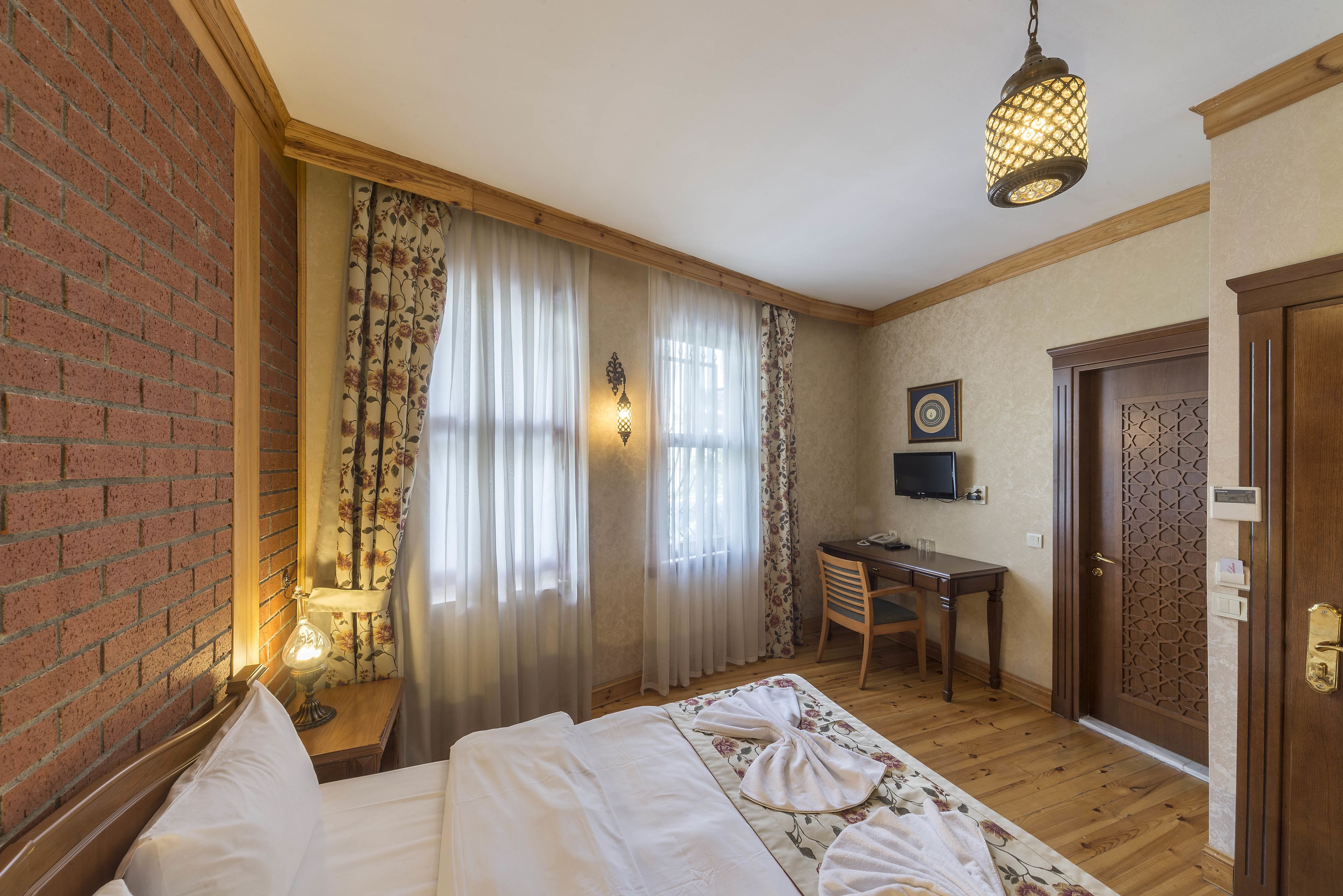A hotel room with a brick wall, a bed with white linens, a desk with a chair, and a wooden door.