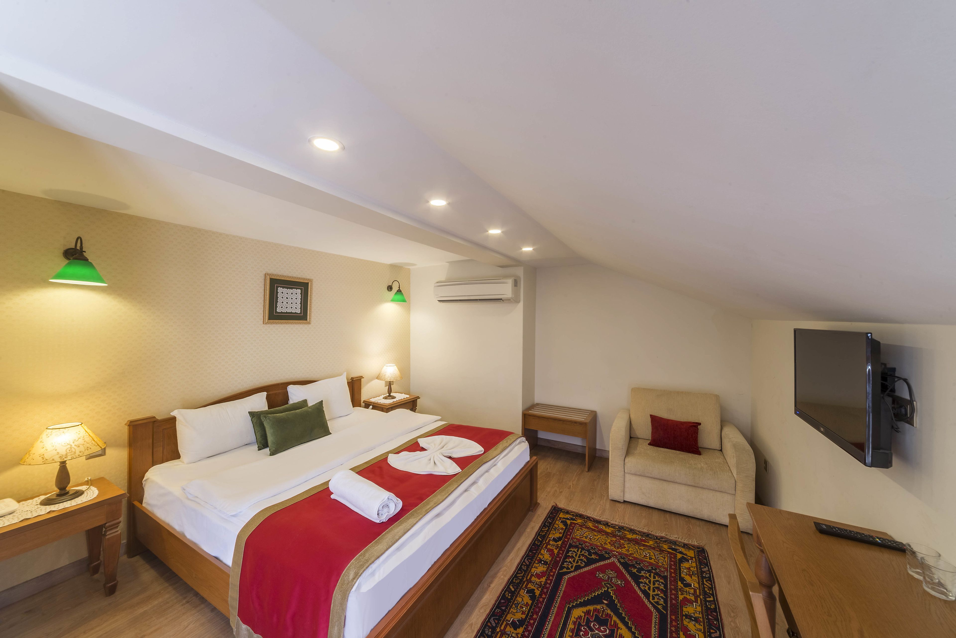 A hotel room with a bed, bedside tables, a chair, a TV, and an air conditioning unit.