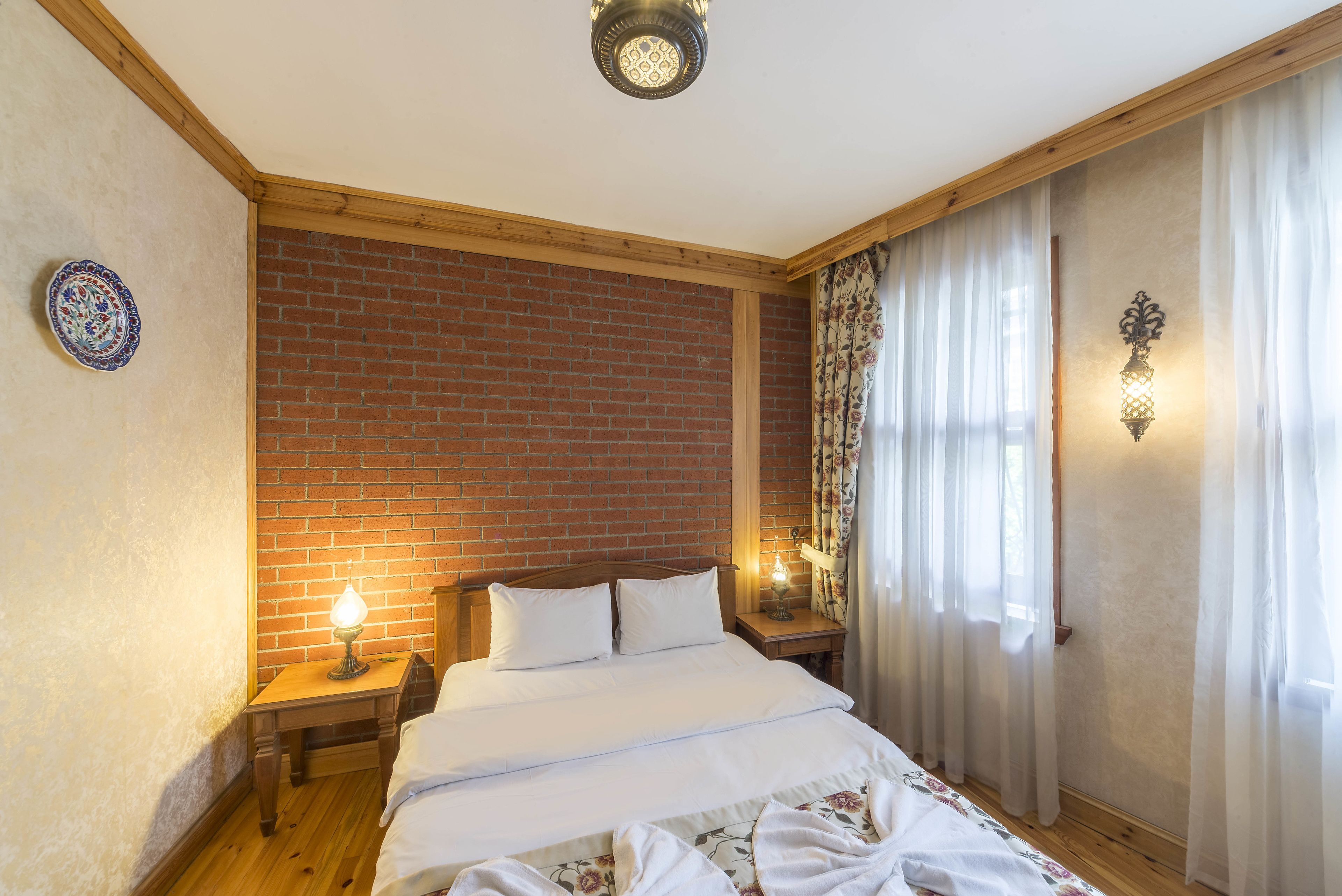 Quarto com parede de tijolos, vigas de madeira no teto, cama com roupa de cama branca, mesa de cabeceira com abajur e um prato decorativo na parede.