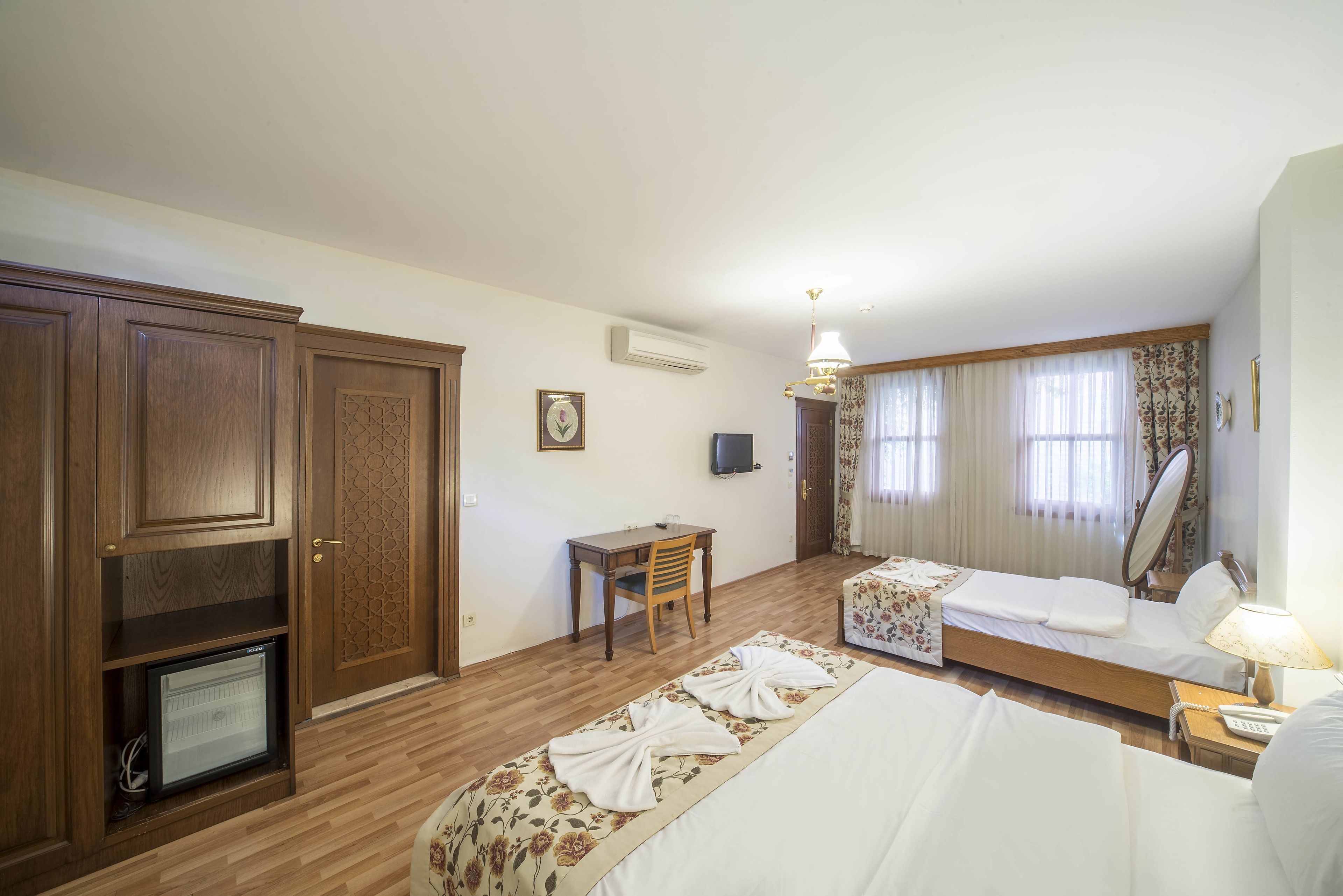 Une chambre d’hôtel avec une armoire en bois, un bureau avec une chaise, un lit recouvert d’une couvre-lit à motifs floraux, une table de chevet avec une lampe et une fenêtre avec des rideaux.