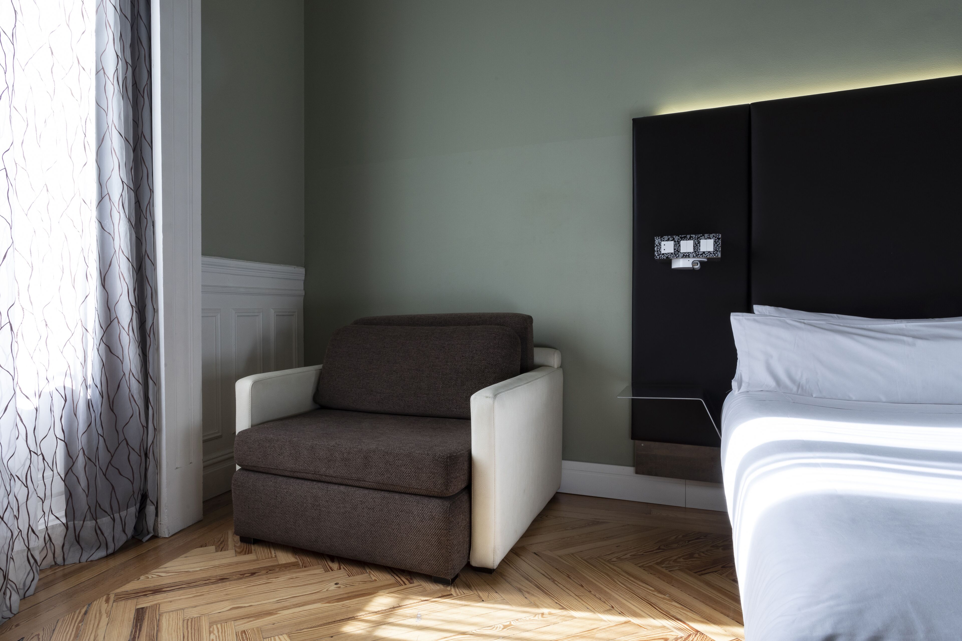 Una habitación de hotel con piso de madera, un sillón marrón, una cama blanca y un cabecero negro.