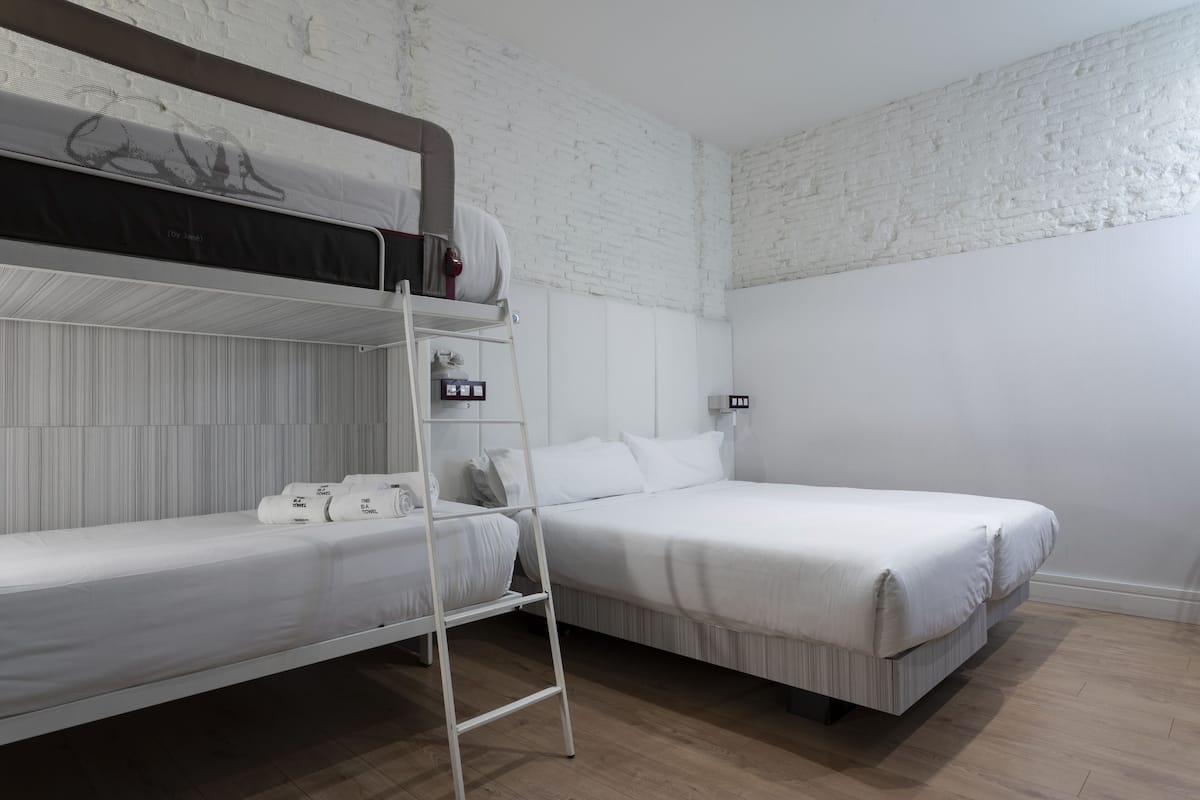Habitación con dos camas, una literas y otra estándar, una escalera y una pared de ladrillo blanco.