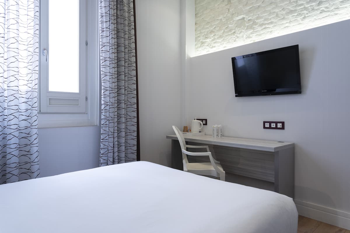 Habitación de hotel con cama, escritorio con silla, televisor de pantalla plana montado en la pared y ventana con cortinas estampadas.