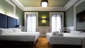 Habitación de hotel con dos camas, piso de madera en espiga, radiador blanco decorativo y televisor de pantalla plana montado en la pared.