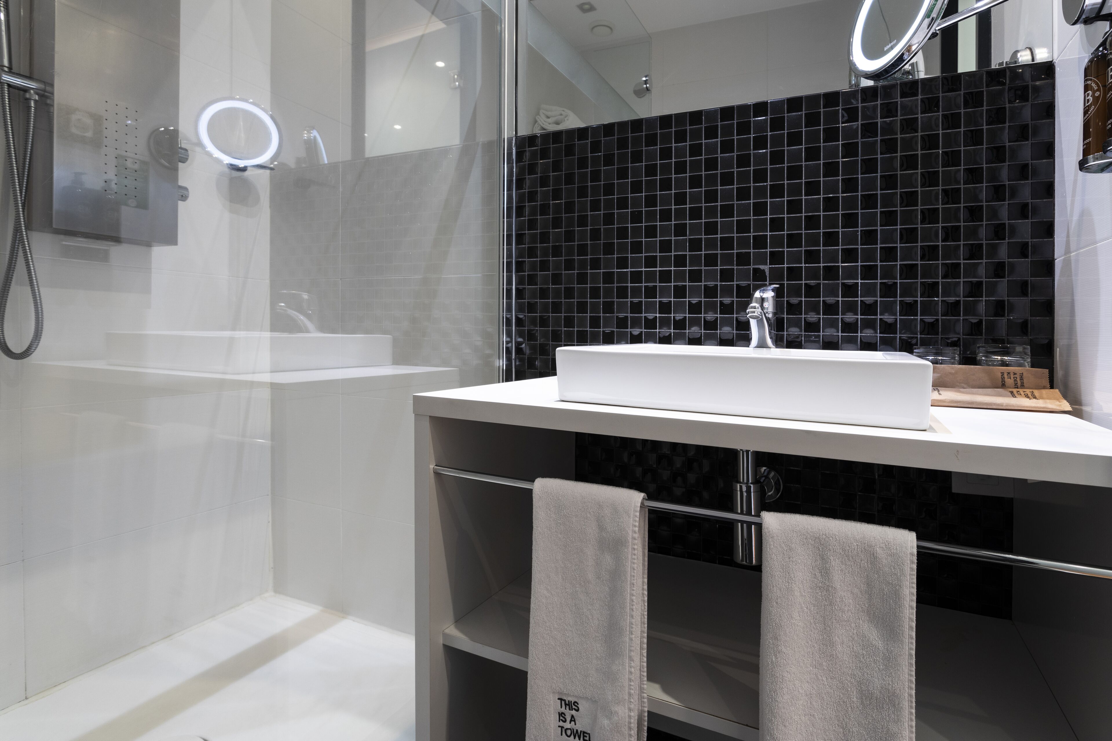 Un baño moderno con ducha envuelta en vidrio, un lavabo blanco y un salpicadero de azulejos negros.