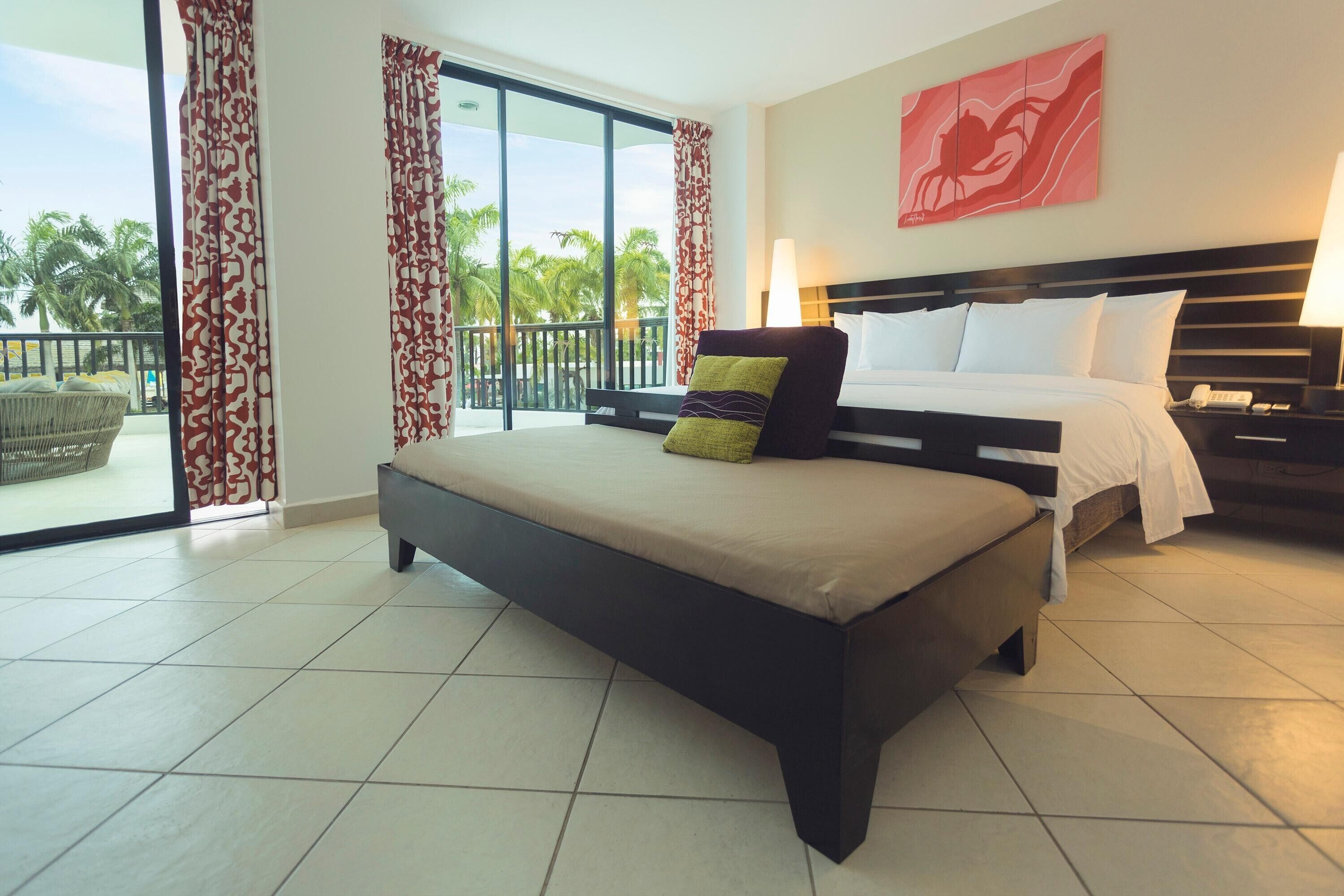 Quarto de hotel com uma cama grande, uma mesa de cabeceira, uma luminária e vista para palmeiras pela varanda.