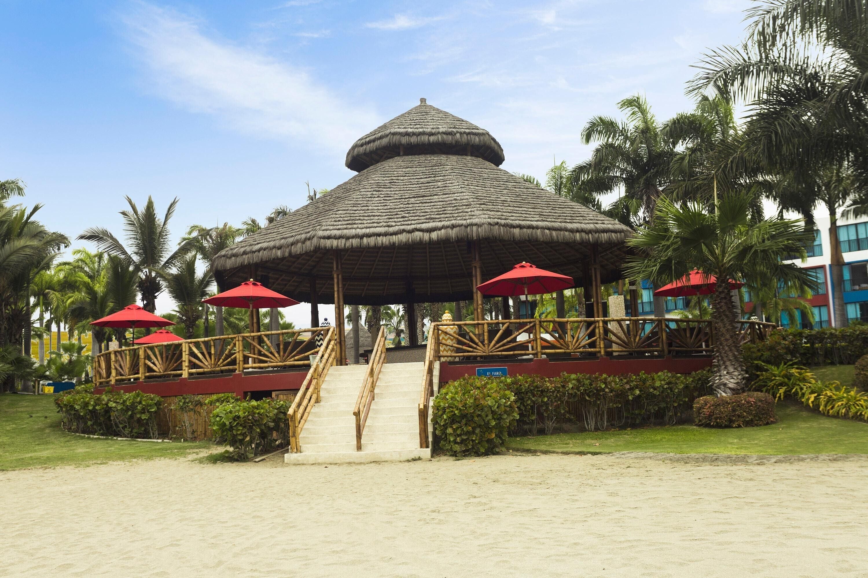 A beachside gazebo with a thatched roof, surrounded by palm trees and a sandy area.