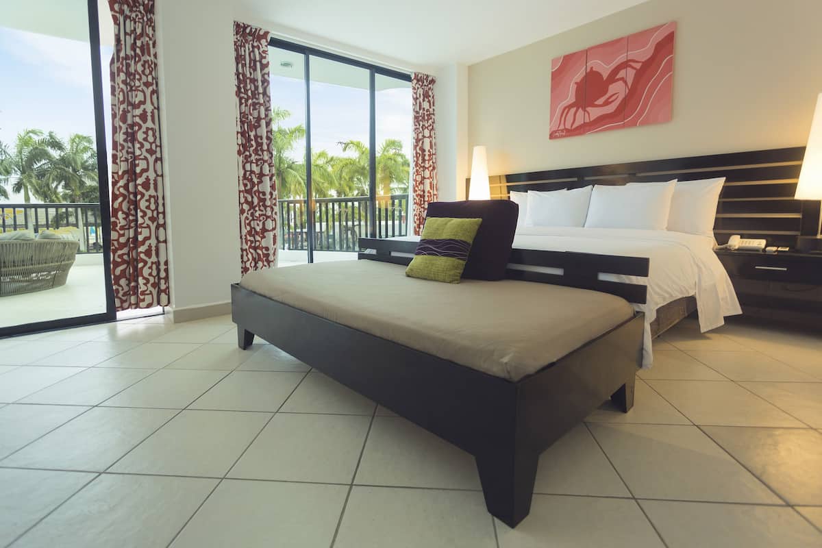 Quarto de hotel com uma cama grande, mesa de cabeceira, luminária e vista para palmeiras através das portas da varanda.