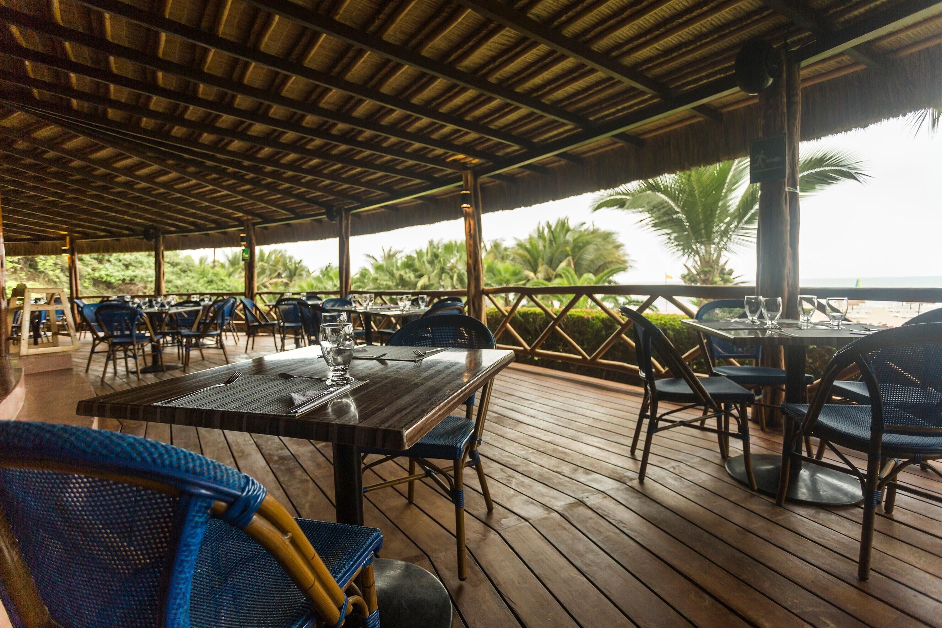 Un espace extérieur aménagé pour les repas, avec des tables et des chaises, une terrasse en bois et une vue sur les palmiers et l’océan.