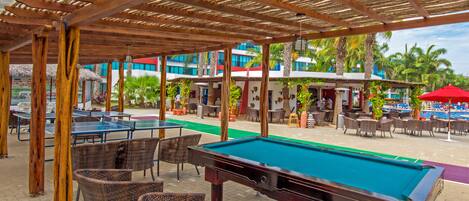 Un espace extérieur avec une piscine, une table de billard, des fauteuils en osier et un coin salon abrité.