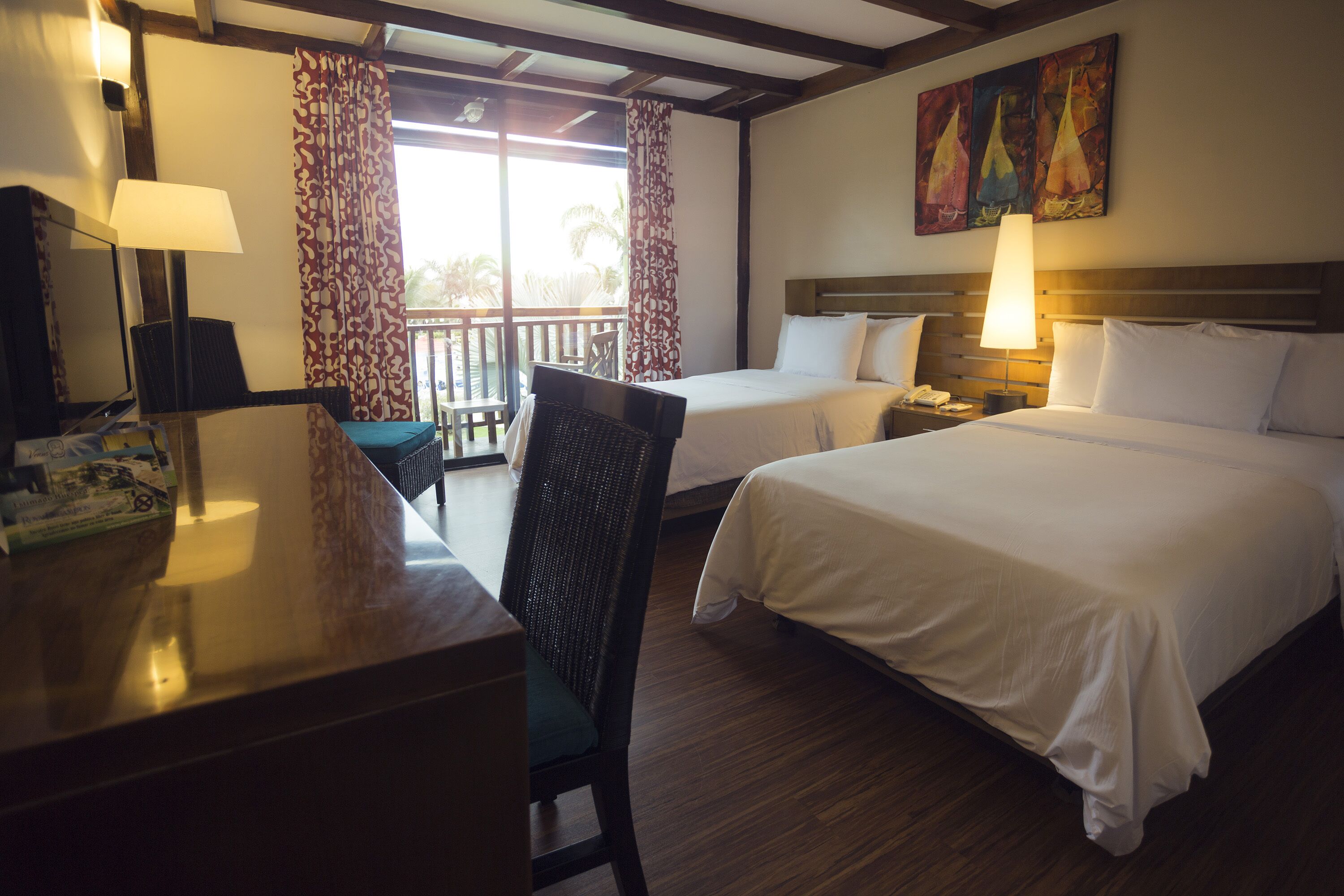 Habitación de hotel con dos camas, un escritorio con lámpara, una silla y un balcón con cortinas.