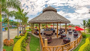 A thatched-roof outdoor bar area with multiple tables and chairs, surrounded by well-maintained landscaping and palm trees. - Royal Decameron Punta Centinela (Santa Elena)