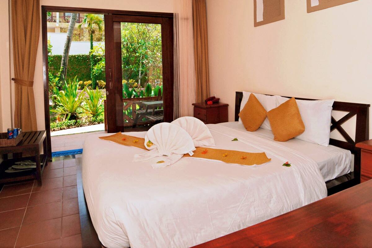 A hotel room with a bed, a wooden nightstand, a small table, and a view of a garden through a sliding door.