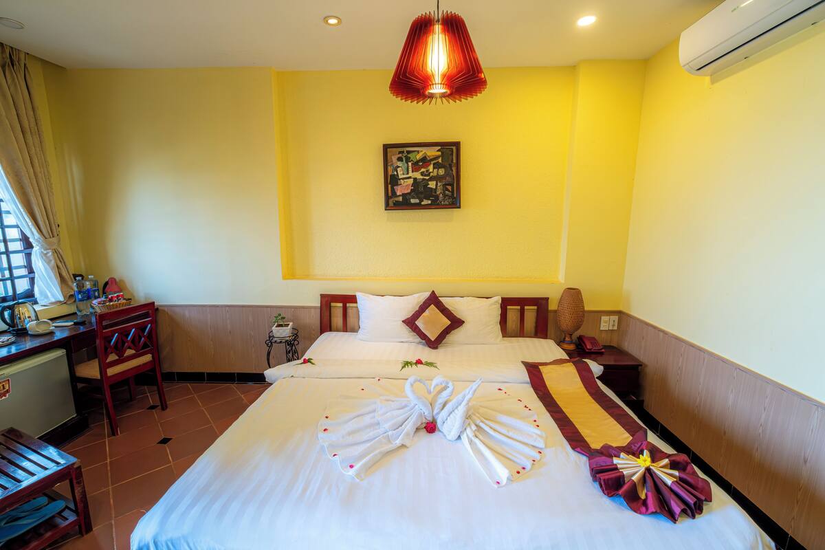 A hotel room with a bed, a desk, a chair, a nightstand, and an air conditioning unit.