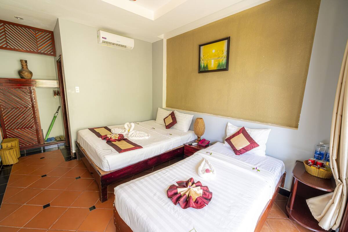 A hotel room with two beds, a wall-mounted air conditioner, a painting, and a small table with a basket.