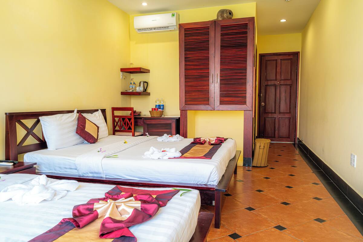 A hotel room with two beds, a wooden wardrobe, a small table with a kettle, and a trash bin.