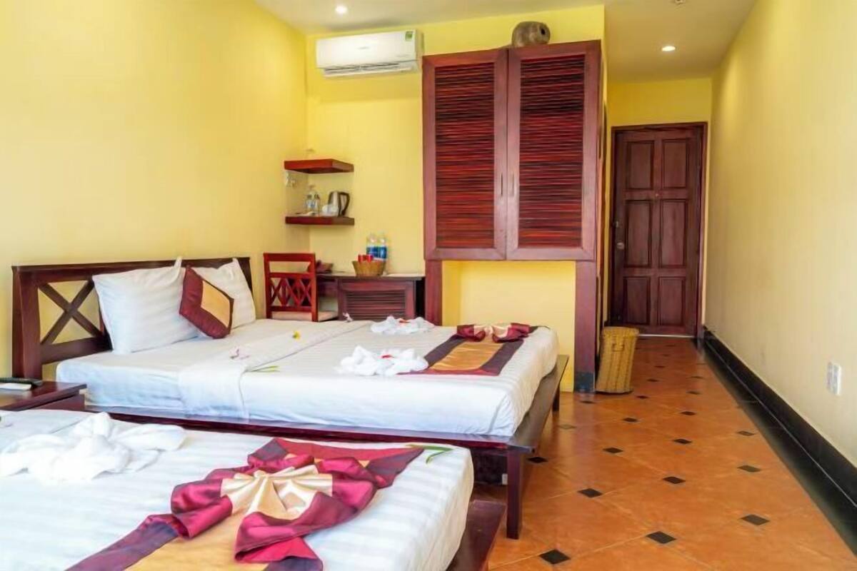 A hotel room with two beds, a wooden wardrobe, a small table with a chair, and a trash bin.