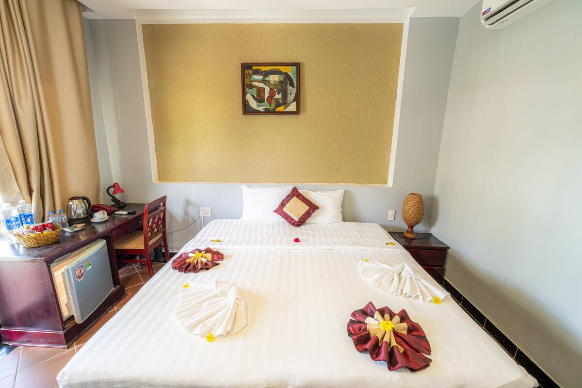 A hotel room with a neatly made bed, a desk with a refrigerator, a lamp, and a wall-mounted painting.