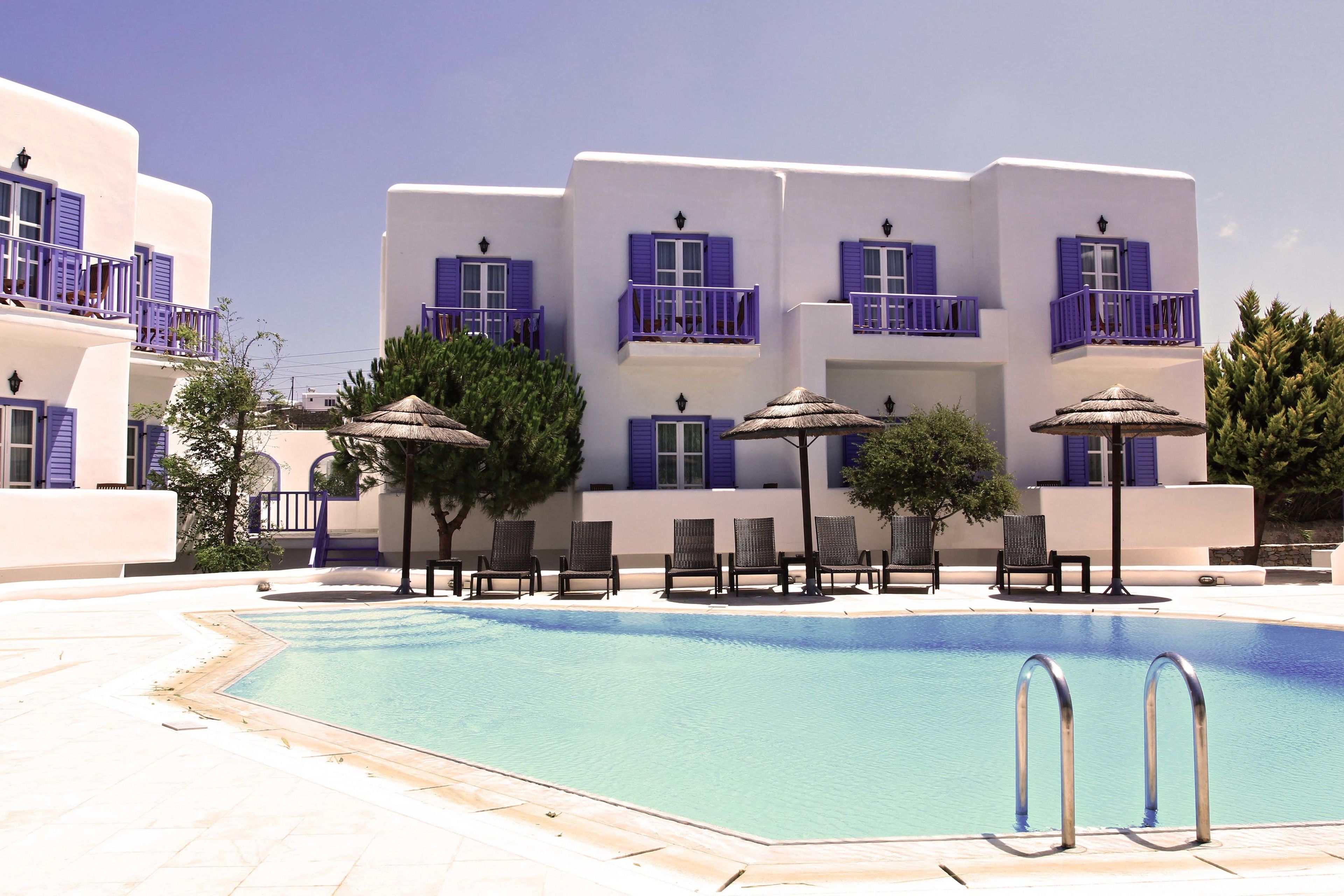 A pool area with a clear blue pool, lounge chairs, and a building with white walls and purple balconies.