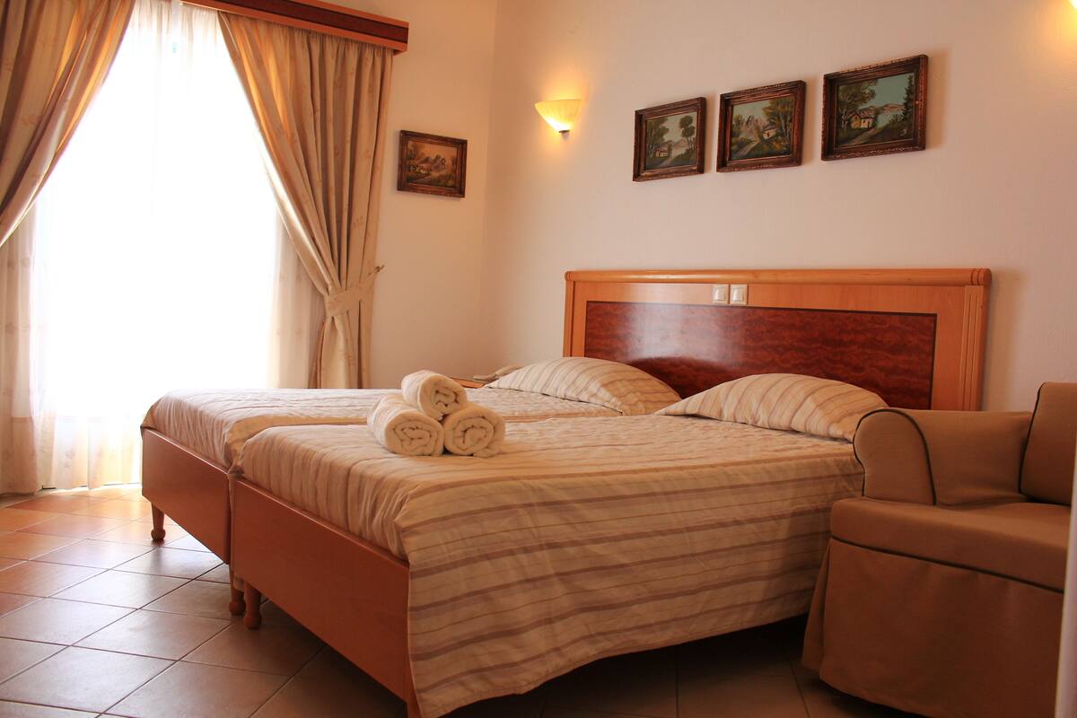 A double bed with a wooden headboard, two pillows, and rolled towels on the bed. There is a small bedside table and a chair to the right. The room has a tiled floor and a window with curtains.
