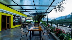An outdoor dining area with tables and chairs, a yellow building, and a view of the mountains. - Westwood Riverside Garden Resort (Devikolam)
