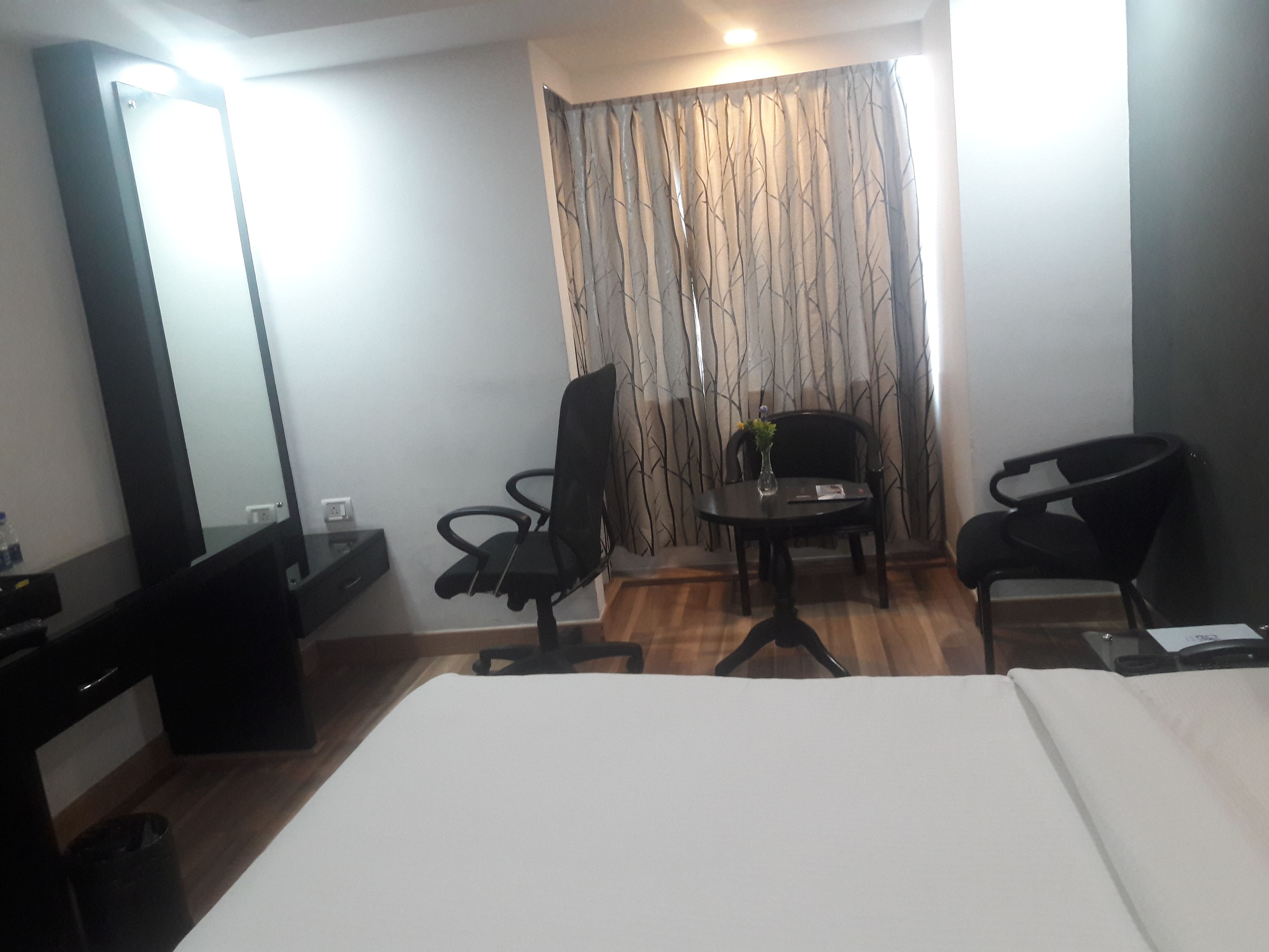 A hotel room with a bed, two chairs, a small table with a vase, a mirror, and a curtain with a tree pattern.