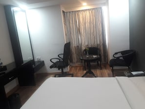 Habitación de hotel con una cama, dos sillas, una mesita con un florero, un espejo y una cortina con estampado de árboles.