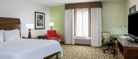 A hotel room with a bed, a desk, a chair, a red armchair, a window with curtains, a wall-mounted TV, and a framed picture on the wall.