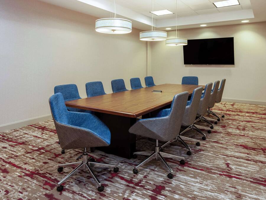 A conference room with a long table, blue and gray chairs, a flat-screen TV on the wall, and patterned carpet.