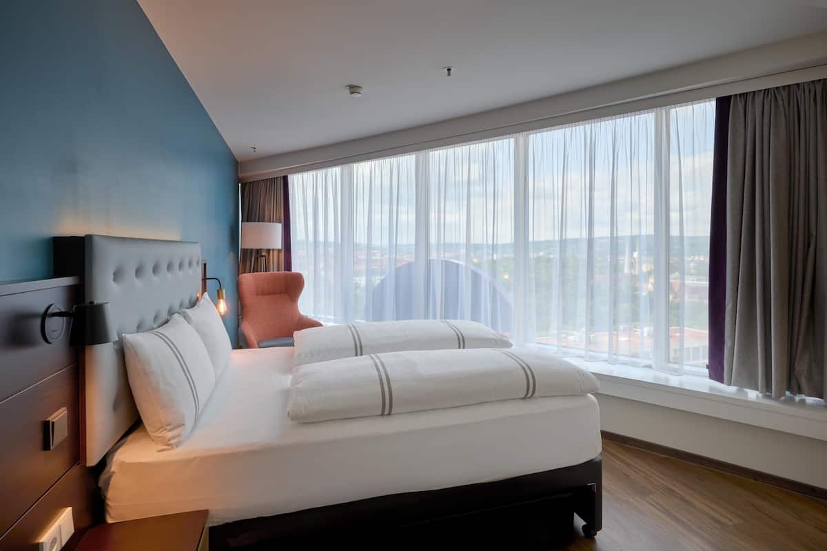 A modern hotel room with a large bed, a headboard with a light fixture, a nightstand, a lamp, and a view of the city through sheer curtains.