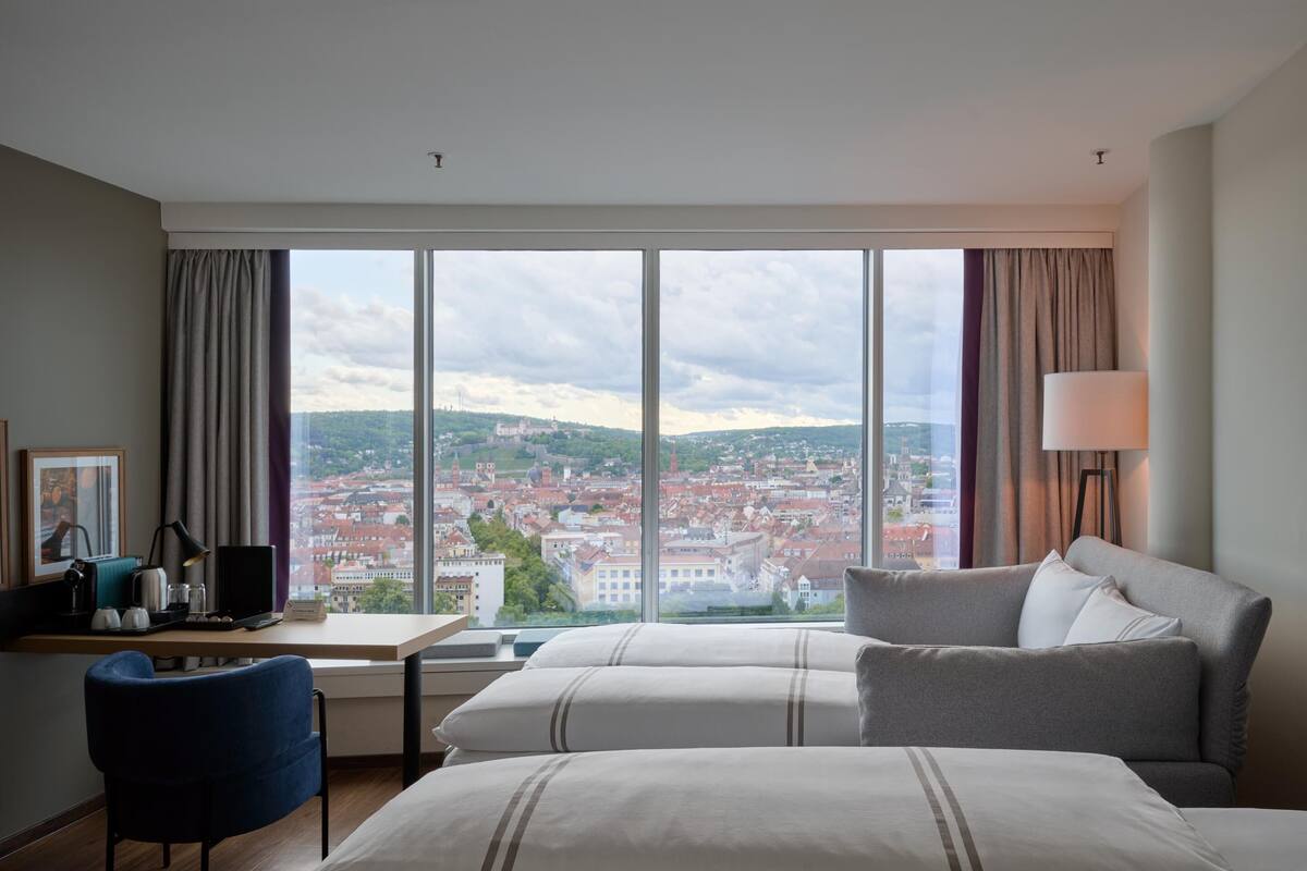 A hotel room with a large window, a desk with a computer, a chair, a bed, and a lamp.