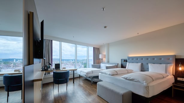 Una habitación de hotel moderna con una cama grande, un escritorio con una silla, un televisor y vistas a la ciudad.