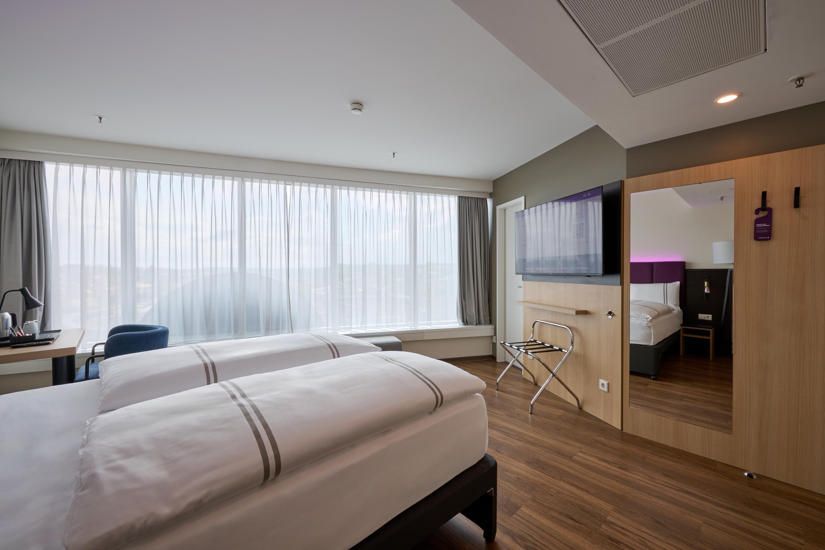 Ein Hotelzimmer mit zwei Betten, einem Schreibtisch mit Stuhl, einem Spiegel und Blick auf die Stadt.