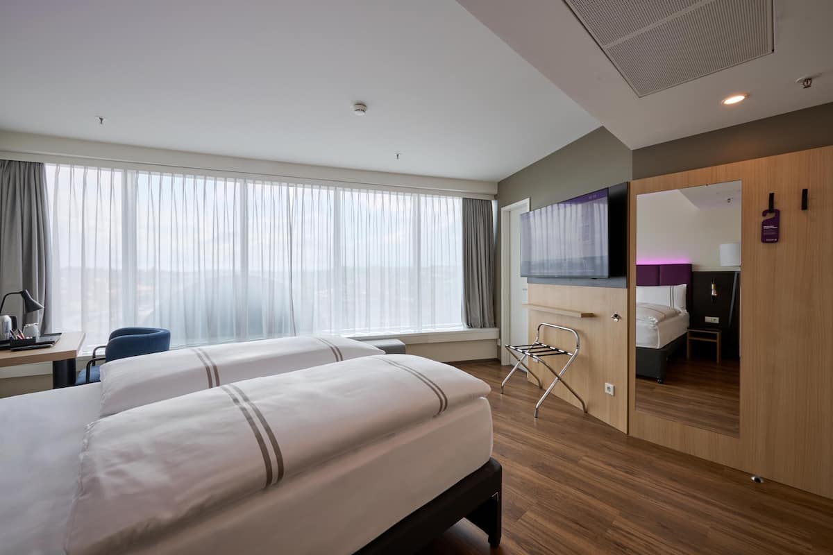 A hotel room with two beds, a desk with a chair, a mirror, and a view of the city.