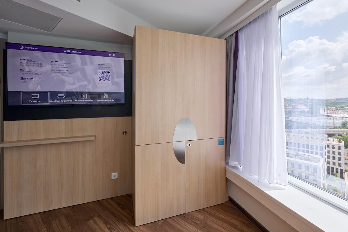 A modern hotel room with a large window, a TV screen displaying hotel information, and a wooden wardrobe.