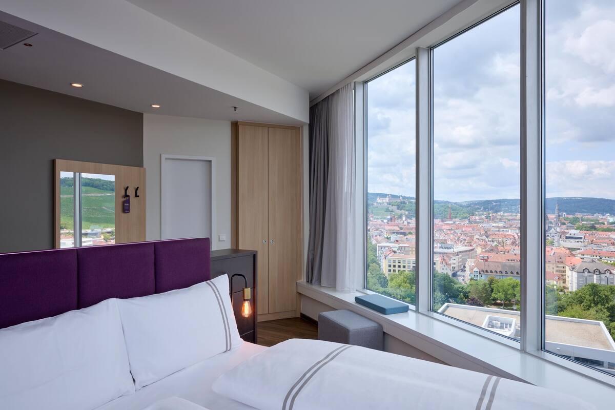 A modern hotel room with a large bed, a wardrobe, a vanity, and a window with a city view.
