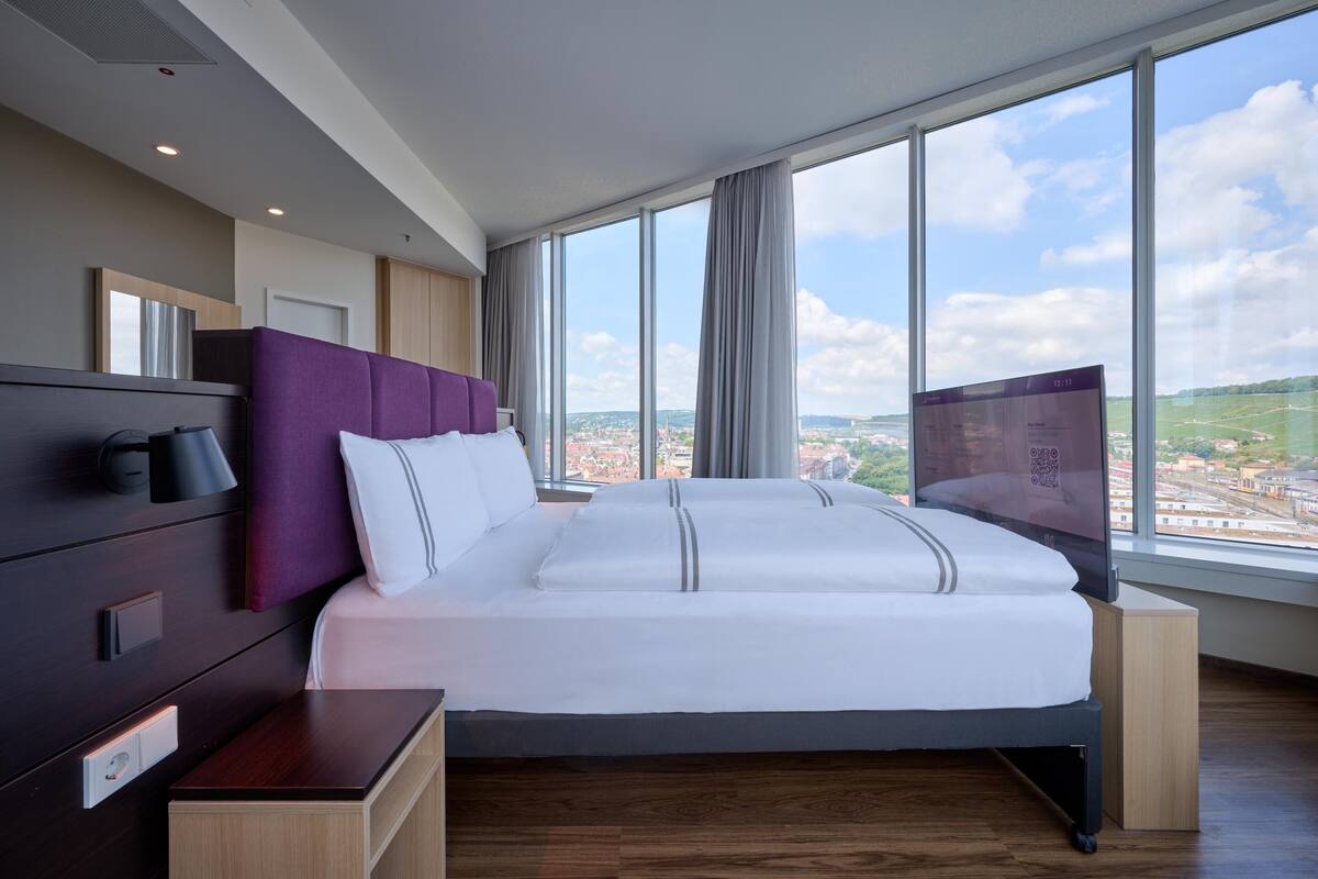 A modern hotel room with a large bed, a TV, a wooden nightstand, and a view of a cityscape through large windows.