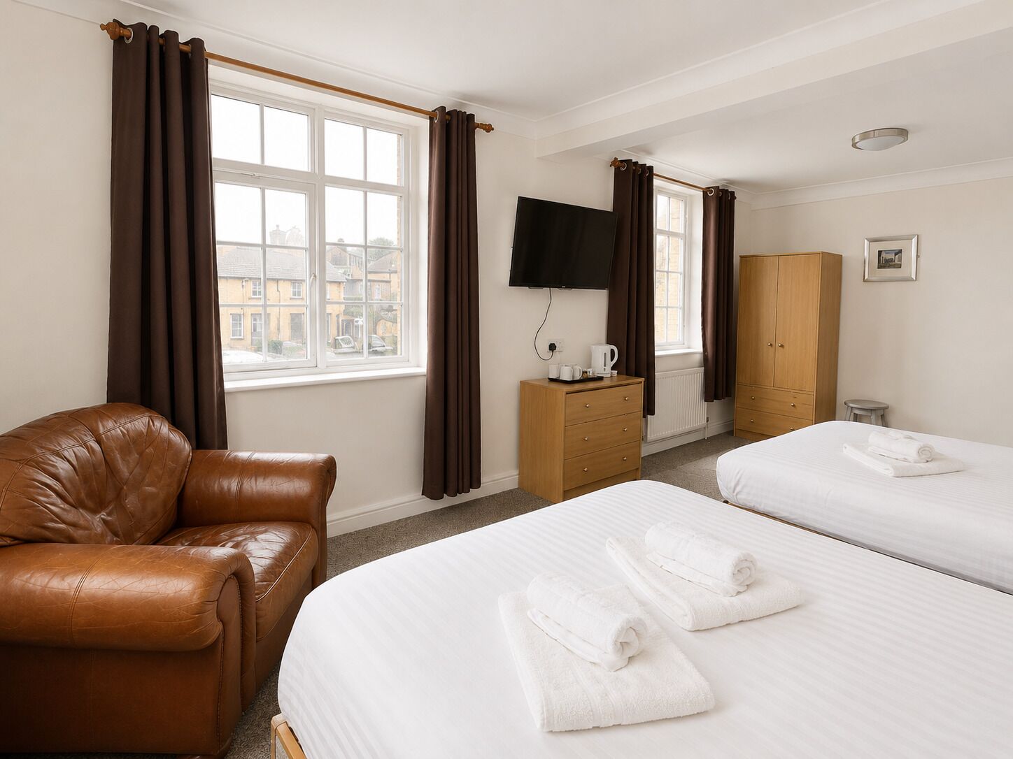 Ein Hotelzimmer mit Ledersessel, zwei Betten mit weißer Bettwäsche, einem hölzernen Schminktisch, einem Fernseher und einem Fenster mit Vorhängen.