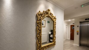 A hallway with a large ornate gold-framed mirror, a radiator, a shelf with folded towels, and an elevator.