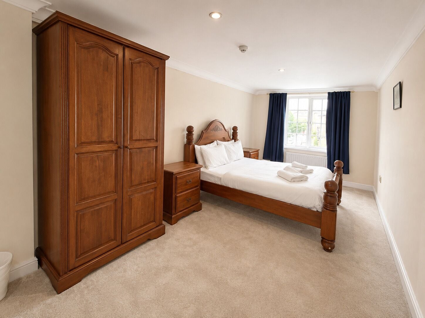 A bedroom with a large wooden wardrobe, a bed with white linens, a nightstand, and a window with curtains.