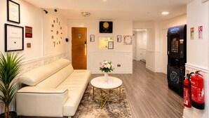 A hotel lobby with a beige sofa, a round table with a vase of flowers, a reception desk, and a water dispenser.