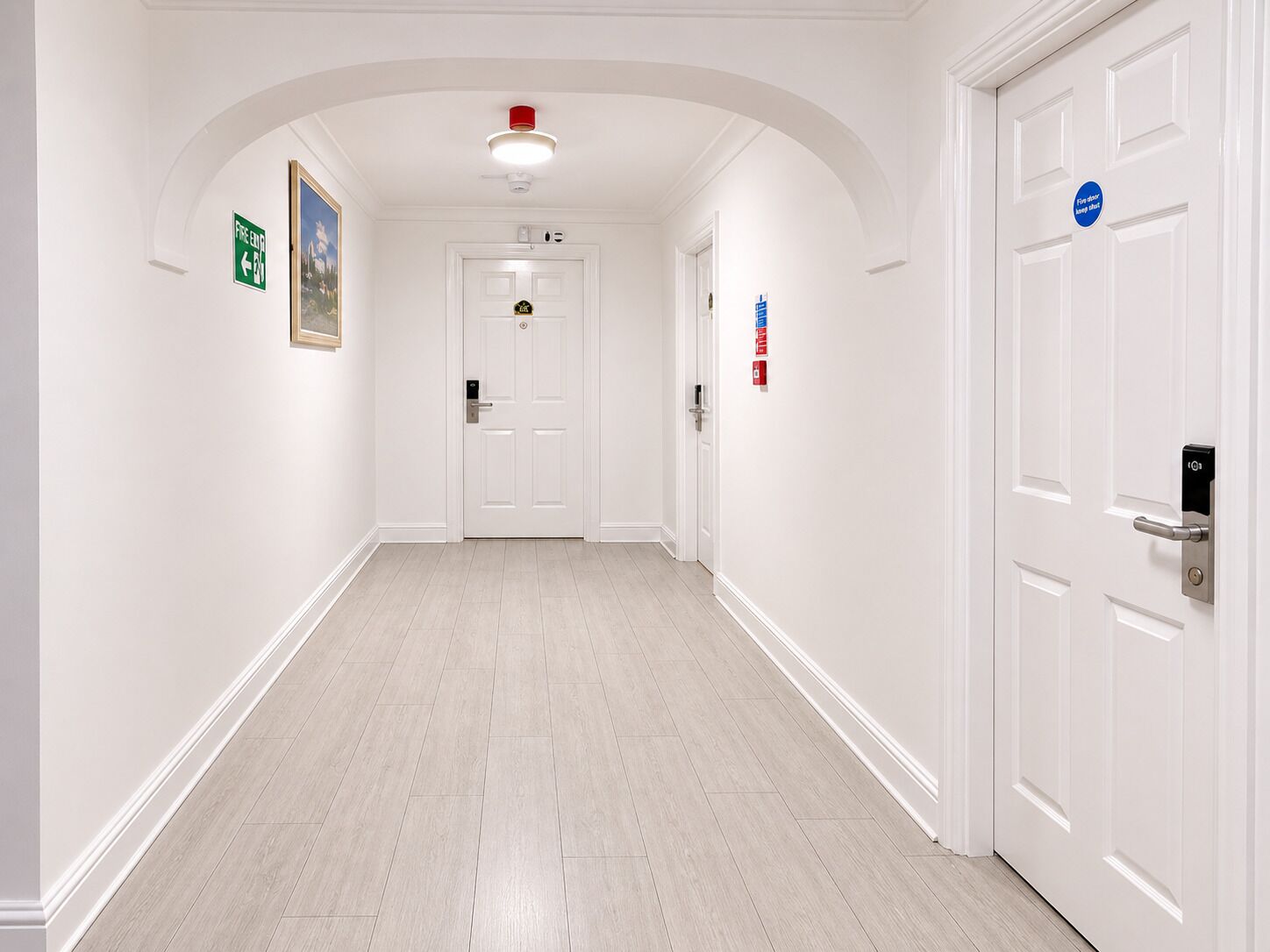A hallway with white walls, a wooden floor, a fire exit sign, and a door with a blue sign.